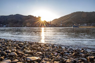 Nehir kenarındaki tepelerde gün batımı. Güneş tepelerin ve üzüm bağlarının üzerinde parlıyor ve nehre yansıyor. Ren 'den Almanya' nın Bacharach kasabasına bakın..