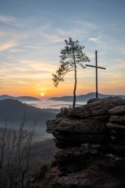 Bir dağ manzarasında gün doğumu. Ormandaki kumtaşı bir uçurumun arkasında yükselen güneş. Almanya 'daki Rtzenfelsen, Palatinate Ormanı' ndaki vadide sis oluşuyor.