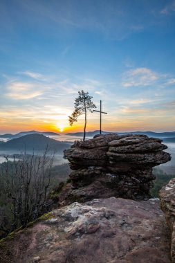 Bir dağ manzarasında gün doğumu. Ormandaki kumtaşı bir uçurumun arkasında yükselen güneş. Almanya 'daki Rtzenfelsen, Palatinate Ormanı' ndaki vadide sis oluşuyor.