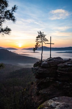 Bir dağ manzarasında gün doğumu. Ormandaki kumtaşı bir uçurumun arkasında yükselen güneş. Almanya 'daki Rtzenfelsen, Palatinate Ormanı' ndaki vadide sis oluşuyor.