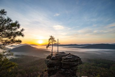 Bir dağ manzarasında gün doğumu. Ormandaki kumtaşı bir uçurumun arkasında yükselen güneş. Almanya 'daki Rtzenfelsen, Palatinate Ormanı' ndaki vadide sis oluşuyor.