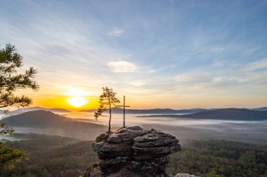 Bir dağ manzarasında gün doğumu. Ormandaki kumtaşı bir uçurumun arkasında yükselen güneş. Almanya 'daki Rtzenfelsen, Palatinate Ormanı' ndaki vadide sis oluşuyor.