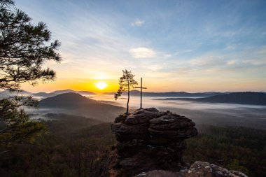 Bir dağ manzarasında gün doğumu. Ormandaki kumtaşı bir uçurumun arkasında yükselen güneş. Almanya 'daki Rtzenfelsen, Palatinate Ormanı' ndaki vadide sis oluşuyor.