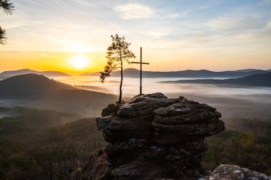 Bir dağ manzarasında gün doğumu. Ormandaki kumtaşı bir uçurumun arkasında yükselen güneş. Almanya 'daki Rtzenfelsen, Palatinate Ormanı' ndaki vadide sis oluşuyor.