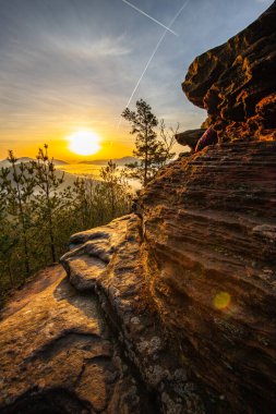 Bir dağ manzarasında gün doğumu. Ormandaki kumtaşı bir uçurumun arkasında yükselen güneş. Almanya 'daki Rtzenfelsen, Palatinate Ormanı' ndaki vadide sis oluşuyor.