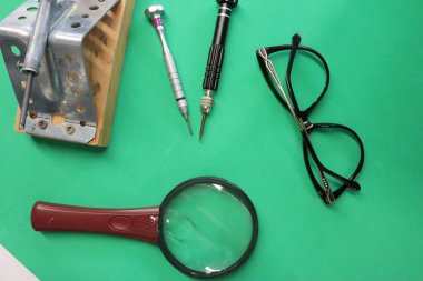 Bir lehim makinesi, bir büyüteç, bir çift gözlük ve bir kalem yeşil bir masa üzerinde sıralanır, genellikle devre bileşeni lehimleme ve donanım programlama için kullanılan araçlar