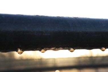 Yakın plan görüntüsü, sabah çiğ damlacıklarının metal bir korkulukta olduğunu ve arka planda çarpıcı bir gündoğumu olduğunu gösteriyor.