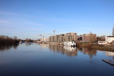 Sahne, nehir kıyısındaki turna, bina ve tekneler ile berrak mavi gökyüzüne kurulmuş kentsel inşaatı yakalıyor.