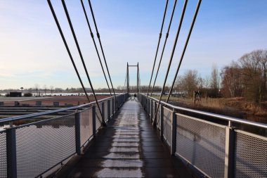 Yapraksız ağaçlar tarafından çerçevelenmiş, berrak kış gökyüzünün altında sakin bir nehir boyunca uzanan bir asma köprü.