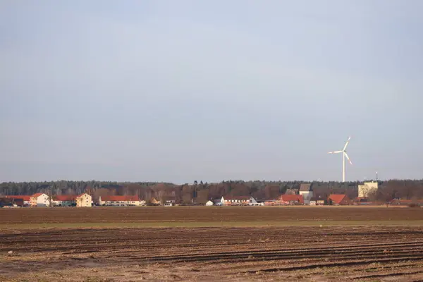 stock image An idyllic rural view with a wind turbine, buildings, and a clear sky, showcasing a serene countryside setting