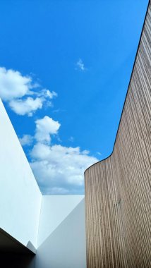 Uzun ve etkileyici bir bina, arkasında yumuşak beyaz bulutlarla süslenmiş canlı bir mavi gökyüzünün altında duruyor.