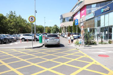 Kalabalık bir alışveriş merkezi otoparkı sayısız araba ve güneş ışığı altında hevesli müşterilerle dolup taşıyor.