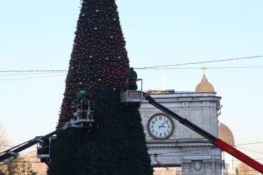 İşçiler kalabalık bir şehir meydanında büyük bir Noel ağacı hazırlıyorlar. Arkasında tarihi bir saat kulesi var.