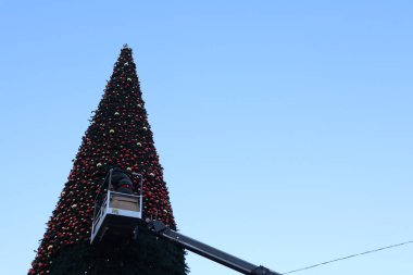 Çalışkan bir işçi, açık mavi gökyüzünün altındaki uzun bir Noel ağacına şenlikli süsler takmak için asansör kullanır.