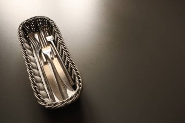 Güzel dokunmuş bir sepet zarif bir şekilde koyu bir yüzeye yerleştirilmiş parlak gümüş çatal bıçak koleksiyonu barındırıyor.