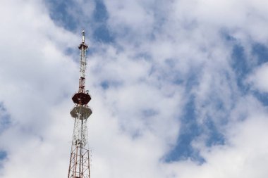 Uzun bir telekomünikasyon kulesi, yumuşak beyaz bulutlarla bezenmiş parlak mavi bir gökyüzüne yükseliyor.