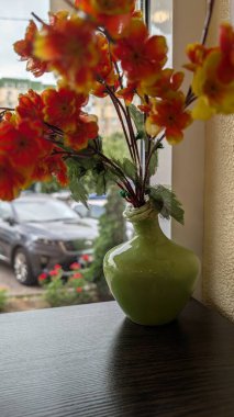 Renkli ve canlı bir suni çiçek sergisi. Bir pencere tarafından yerleştirilmiş zarif yeşil bir vazoya yerleştirilmiş.