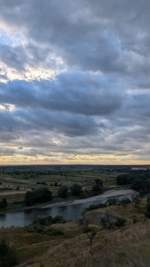 Sersemletici bir manzara, çarpıcı bulutlu gökyüzünün altında yemyeşil çayırlarda akan dolambaçlı bir nehri gösteriyor.