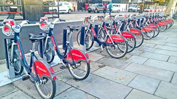 Santander cycles in a diagonal row on docking station at London Bridge Trotoar, with bus stop and car parking in the background.