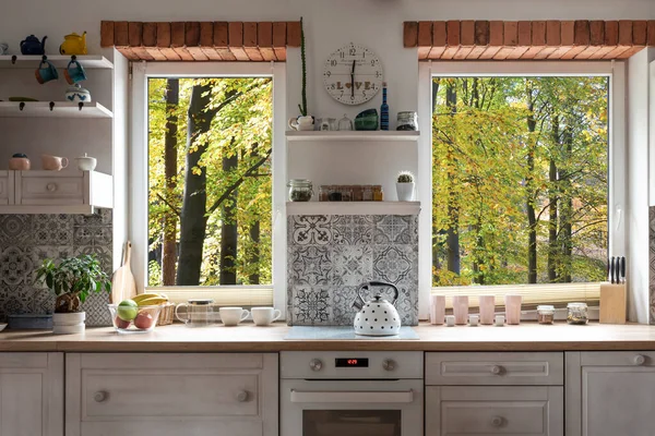 Interior of kitchen with view from window on forest, white wooden furniture and bricks as a decoration. Scandinavian style in a cottage with countertop and modern tiles. Stylish architecture.