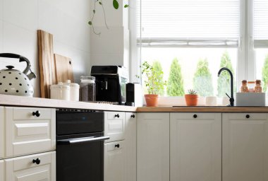 Pencerenin yanında musluk, perdeler ve ahşap tezgahta yeşil bitkiler. Çekmeceleri ve dolapları olan beyaz mobilyalar ve modern mutfak içinde çaydanlığı olan fırın.. 