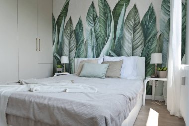 Yatak odasının dekorasyonu, penceresi ve duvarında yeşil yapraklar olan bir duvar kağıdı. Battaniye ve yastıkla rahat bir yatak..