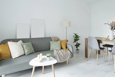 Oturma odasının içinde yastıklı rahat kanepe, sandalyeli masa ve beyaz ahşap zemin. Beyaz duvardaki gibi boş bir çerçeve..