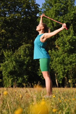 Genç bir kadın parkta lastik bantla poz veriyor ve vücut egzersizi yapıyor. Jimnastik yaparken spor giyim sektöründe aktif bir kız. Dikey.