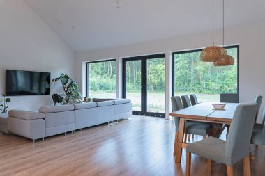 Yeni evinde penceresi olan modern bir oturma odası. İçi yemek masası, ahşap zemin, rahat kanepe ve duvarda televizyon.. 