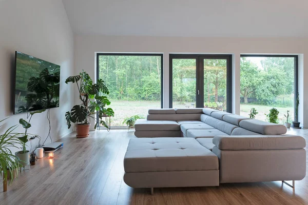 Comfortable sofa, tv on the wall, wooden floor and large window in modern interior of living room at new home.