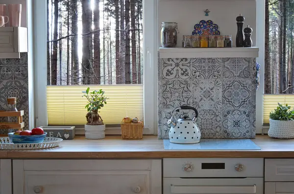 Interior of kitchen with view from window on forest, design pattern tiles on the wall, white wooden furniture, oven and kettle on the counter. Scandinavian cottage with pattern tiles on the wall