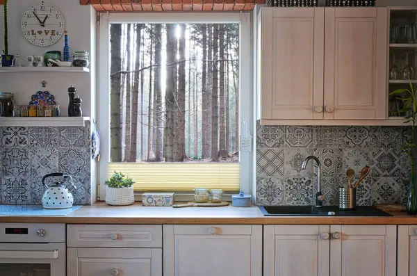 Interior of kitchen with view from window on forest, design pattern tiles on the wall, white wooden furniture, oven and kettle on the counter. Scandinavian cottage with pattern tiles on the wall