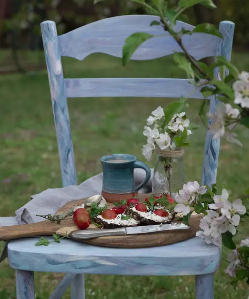 Food for breakfast on the rural chair in the natural green garden in the spring. Beautiful blooming flowers with healthy meal on a wooden board. Fresh vegetable, sandwich and coffee. Outdoor.
