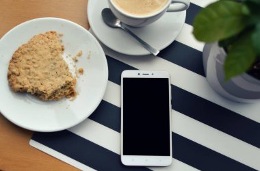 Siyah ve beyaz çizgili arka planda bir tabakta bir fincan kahve ve kek ile boş ekranlı akıllı telefon. Masa üstü görünümü. Öğle arasında iş ve teknoloji arasında. Model.