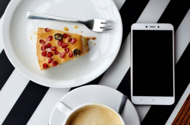 Siyah ve beyaz çizgili arka planda bir tabakta bir fincan kahve ve kek ile boş ekranlı akıllı telefon. Masa üstü görünümü. Öğle arasında iş ve teknoloji arasında. Model.
