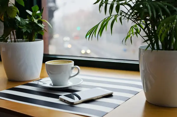 Cup of coffee, smartphone and plants on wooden table and striped background near window in cafe. Relax time and break.