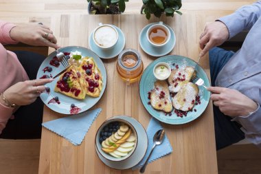 İnsanların elinde çatal bıçak takımı olan bir tabakta kahvaltı için farklı renkte yemekler. Sağlıklı yiyecekler, restorandaki ahşap masada meyveli lapa ve krepli krepler. Üst manzara düzdü.