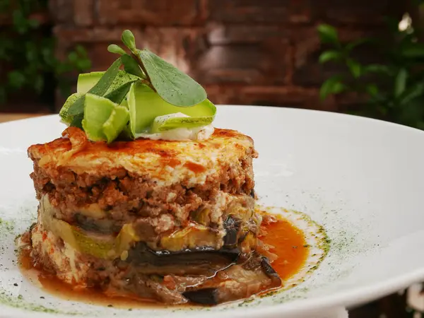 Moussaka as a traditional greek dish for dinner on a white plate in restaurant. Meal made from aubergine and meat in baking form. Ready for eat. Close up.