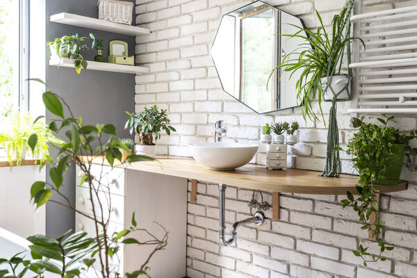 Interior of bathroom in industrial style with white bricks on the wall, stylish mirror, green plants, wooden counter with ceramic sink. Spa with plant and window.