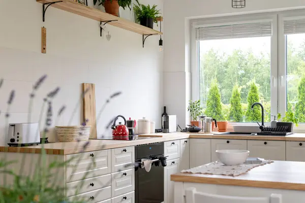 stock image Modern interior of white kitchen with window in rural style. Wooden furniture, countertop, stove and plants in design kitchen. Stylish interior.