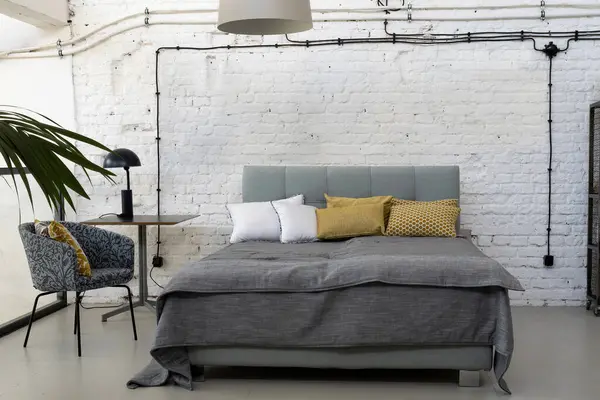 Industrial interior of bedroom in scandinavian style with grey bed, pillows and white brick wall with copy space. Indoors architecture design.