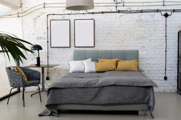 Industrial interior of bedroom in scandinavian style with grey bed, pillows and white brick wall with blank picture frame with mock up. Indoors architecture design.
