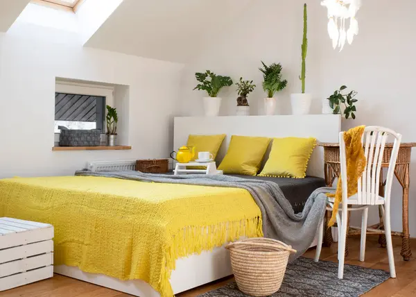 Boho bedroom in the attic with bed with yellow blanket and pillows. Stylish white interior with yellow decoration at home.