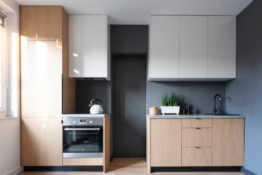 Beyaz mobilyaları, ahşap tarzı dolabı ve gri duvarı olan modern mutfak. Minimalist tarzda rahat bir daire. Yeni çağdaş ev