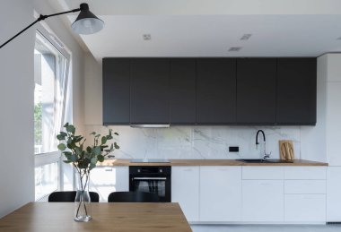 Duvarında mermer ve sandalyeli yemek masası olan lüks bir mutfak. Pencereli yeni dairede beyaz mobilyalar ve ahşap tezgah.