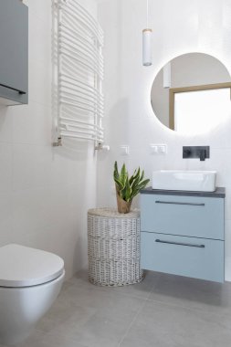 Luxurious and interior of bathroom with ceramic washbasin on the cabinet, round mirror o the white wall with led lights and toilet. Vertical.