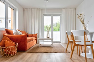 Modern living room with red sofa with pillows, stylish table with chairs and wooden floor. Cozy interior of room with big window and design decor in new apartment in hotel. 