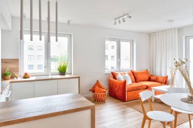 Living room interior in new apartment with modern red sofa with pillows, window, wooden floor and table with chairs. Luxury and minimalism at cozy home.