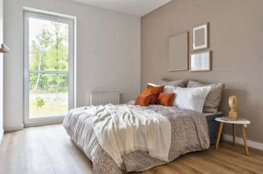 Comfortable double bed with red piloows, white bedding and blanket in cozy bedroom. Stylish interior in hotel with frame on the wall. Modern room in apartment at home with window.