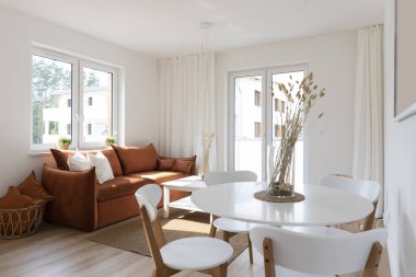 Modern living room with red sofa with pillows, stylish table with chairs and wooden floor. Cozy interior of room with big window and design decor in new apartment in hotel.